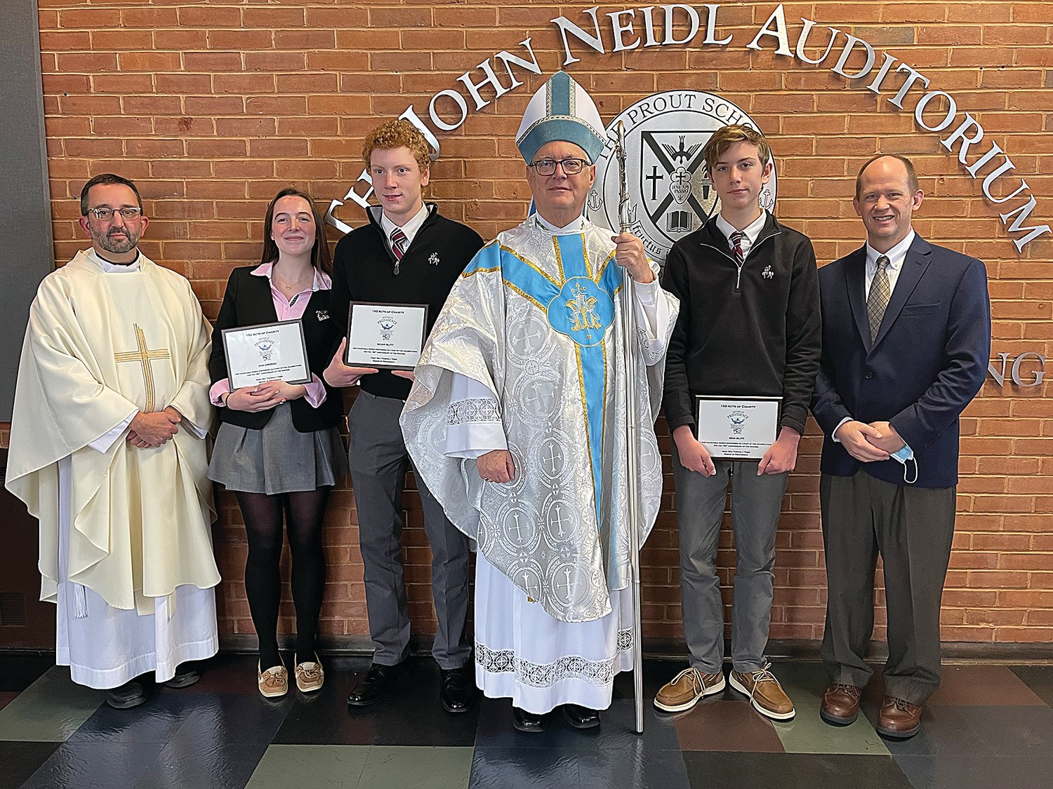 Bishop Thomas J. Tobin presents the first of the 150 Acts of Charity Awards to Prout School students Ava Grosso and brothers Noah and Ben Slitt. Prout Chaplain Father Carl Fisette and Secretary of Catholic Charities and Social Ministry James Jahnz join the group for a photo.